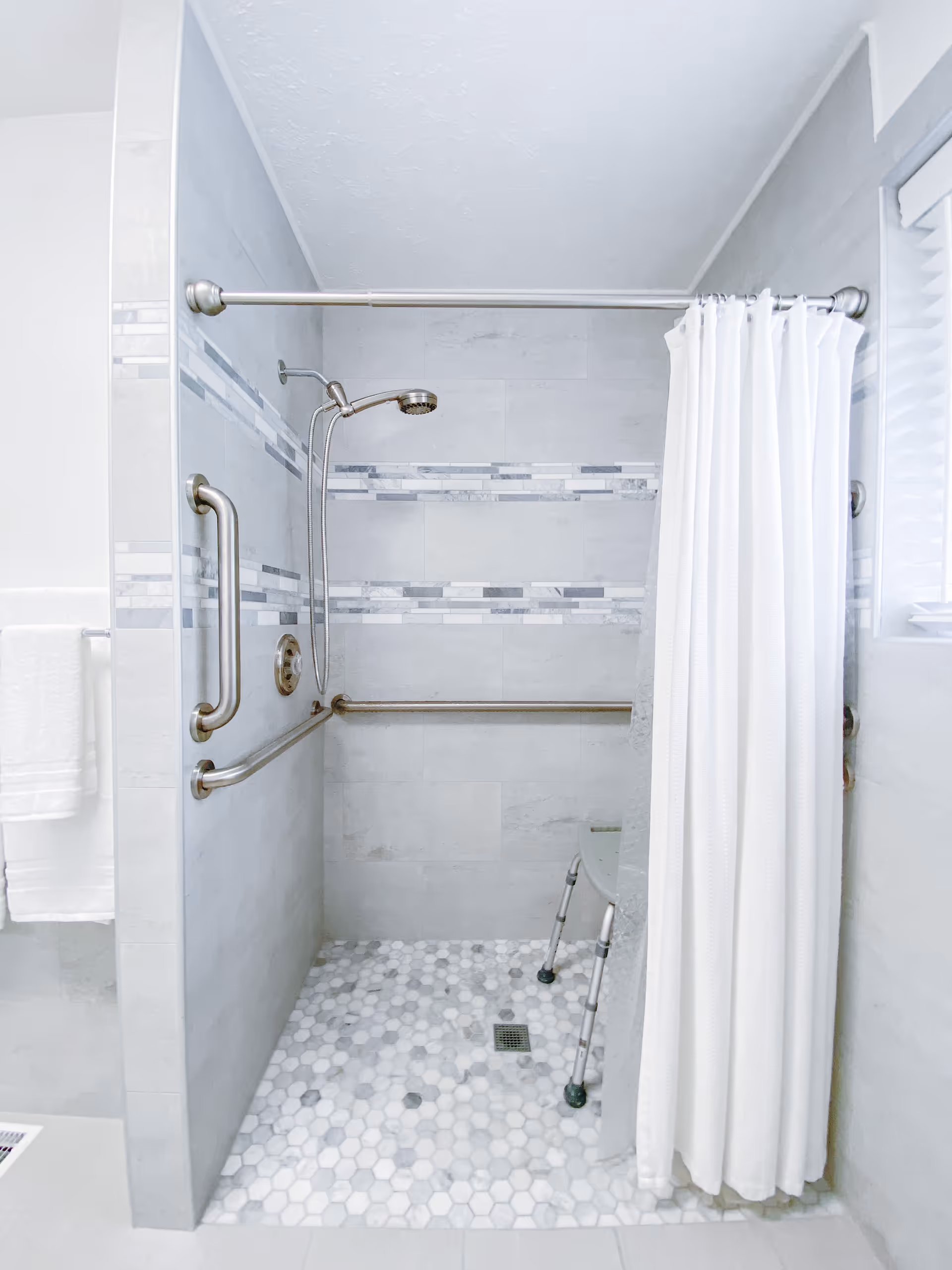 A modern walk-in shower with light gray tiles and decorative horizontal tile accents. The shower has a handheld showerhead, stainless steel grab bars on the walls, a white shower curtain partially drawn, and a small shower chair inside. The floor is covered with hexagonal mosaic tiles in shades of white and gray.