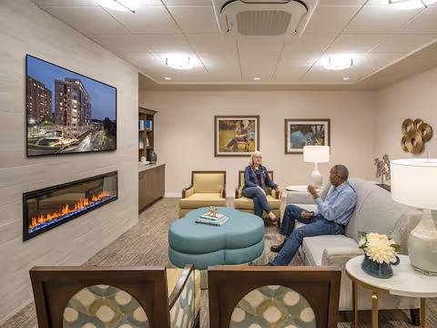A cozy living room in a senior living facility with two people sitting and talking. The room features a modern electric fireplace, a large wall-mounted TV displaying a building exterior, comfortable seating including a sofa, armchairs, and a round ottoman. The walls are decorated with framed artwork and the room is softly lit with ceiling lights and table lamps.