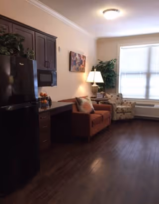 Interior view of a senior living facility room featuring a black refrigerator, dark wood cabinets, a microwave, a small desk area, a brown sofa with pillows, a floral armchair, a floor lamp, a large window with blinds, and a potted plant in the corner.