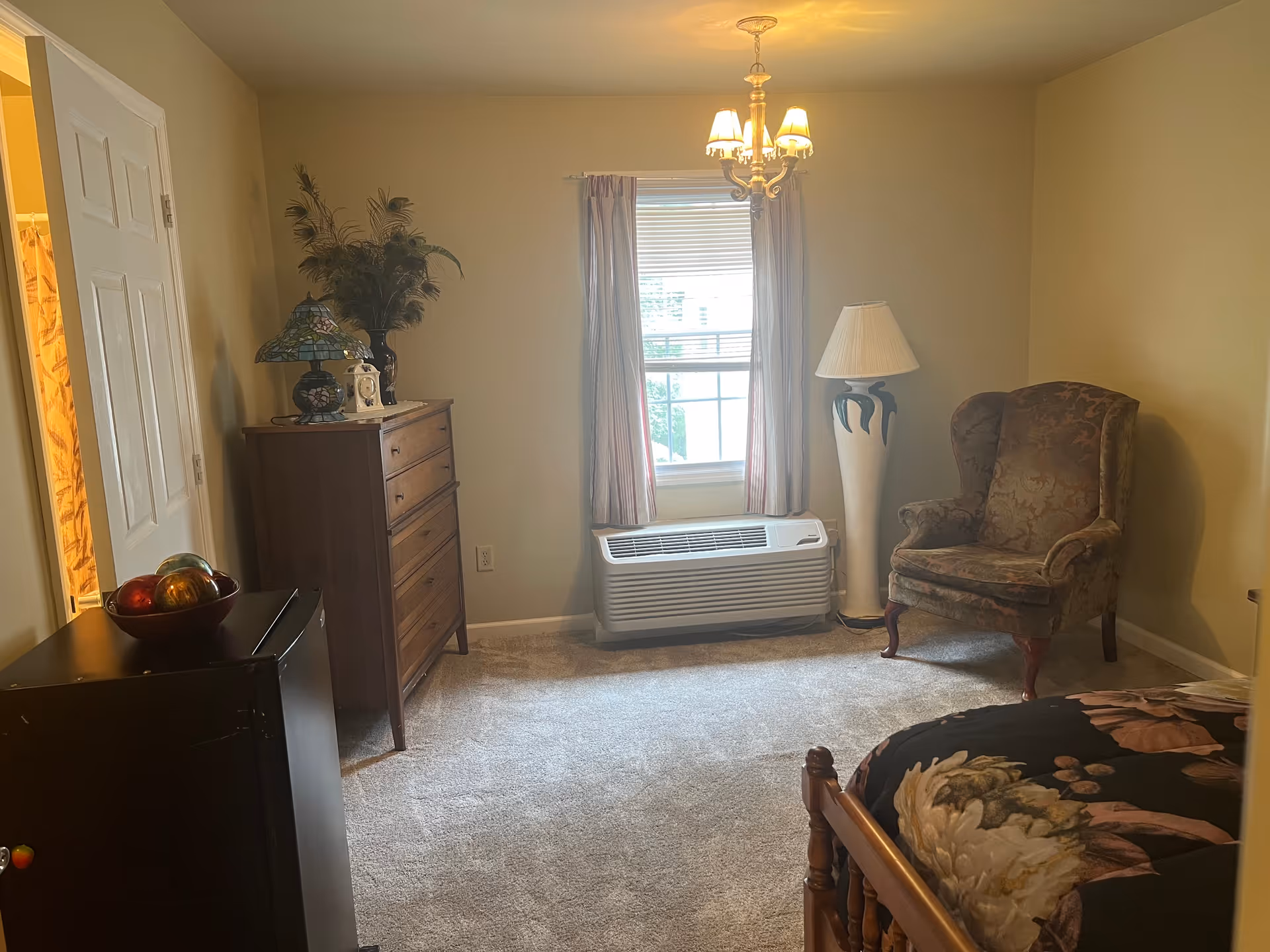 A cozy bedroom with beige walls and carpeted floor. The room features a wooden bed with floral bedding, a vintage upholstered armchair, a tall wooden dresser with decorative items on top, a small black refrigerator with a bowl of decorative balls, a window with curtains and a heating/cooling unit beneath it, and a floor lamp with a white shade. A chandelier with three lights hangs from the ceiling.