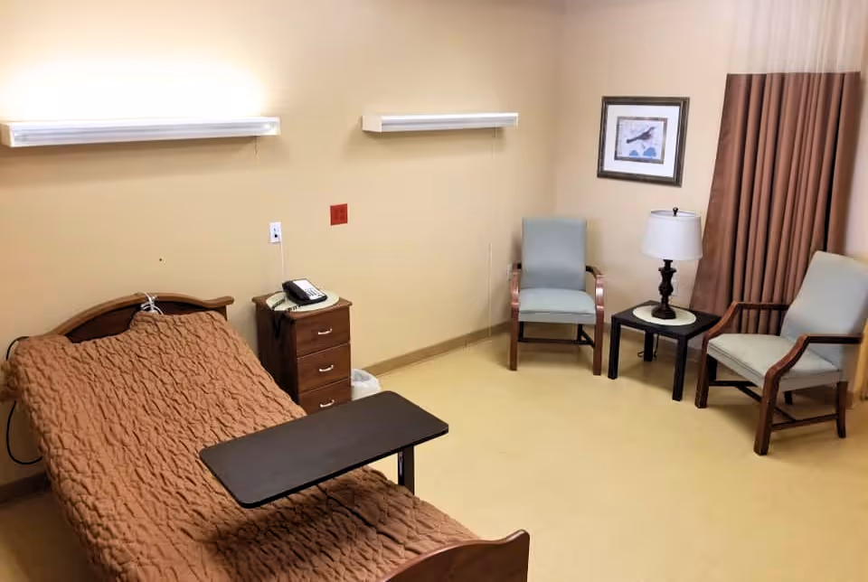 A nursing facility room with a single bed covered in a brown quilt, a small bedside table with a telephone, two light fixtures on the wall above the bed, two armchairs, a small round table with a lamp, a framed picture on the wall, and brown curtains covering a window.