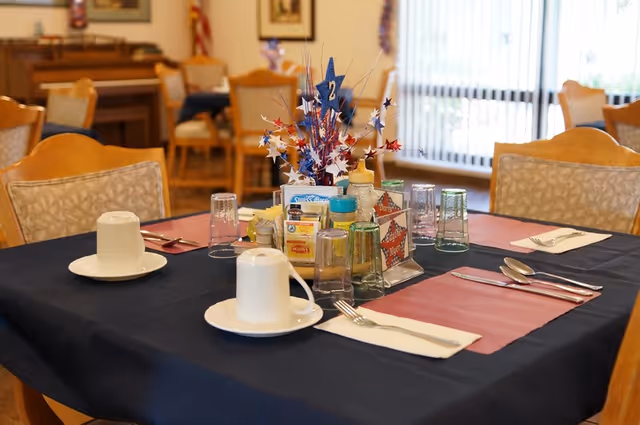 A dining table set for a meal with cups, glasses, utensils, and condiments in a senior living facility dining room. The table has a dark blue tablecloth with red placemats and a patriotic centerpiece with red, white, and blue star decorations. Chairs and other tables are visible in the background along with a piano and framed pictures on the wall.