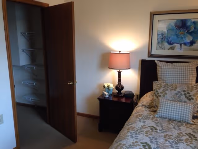 A bedroom with a bed on the right side featuring floral and checkered pillows and bedding. Next to the bed is a dark wooden nightstand with a lamp, a small flower arrangement, and an alarm clock. A framed picture of a blue flower hangs on the wall above the bed. To the left, there is an open door leading to a closet with wire shelving.