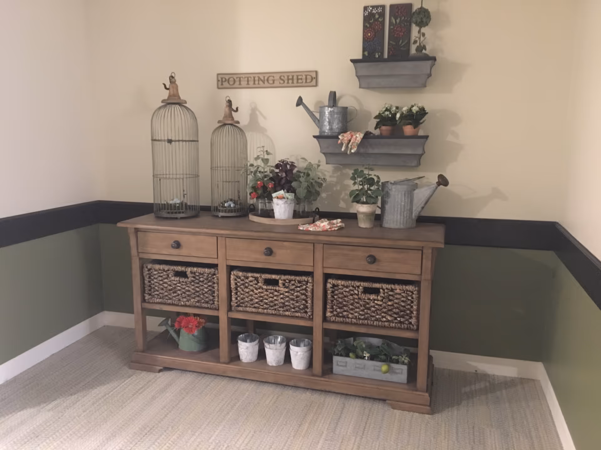 Wooden console table with wicker baskets and gardening decor including potted plants, watering cans and a 'POTTING SHED' sign on a beige and green wall.