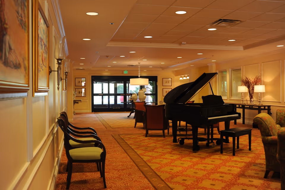 A spacious, warmly lit common area with a grand piano, several chairs, and sofas. The room features patterned carpet, framed artwork on the walls, and large windows letting in natural light. There are also decorative lamps and vases on tables, creating a cozy and inviting atmosphere.