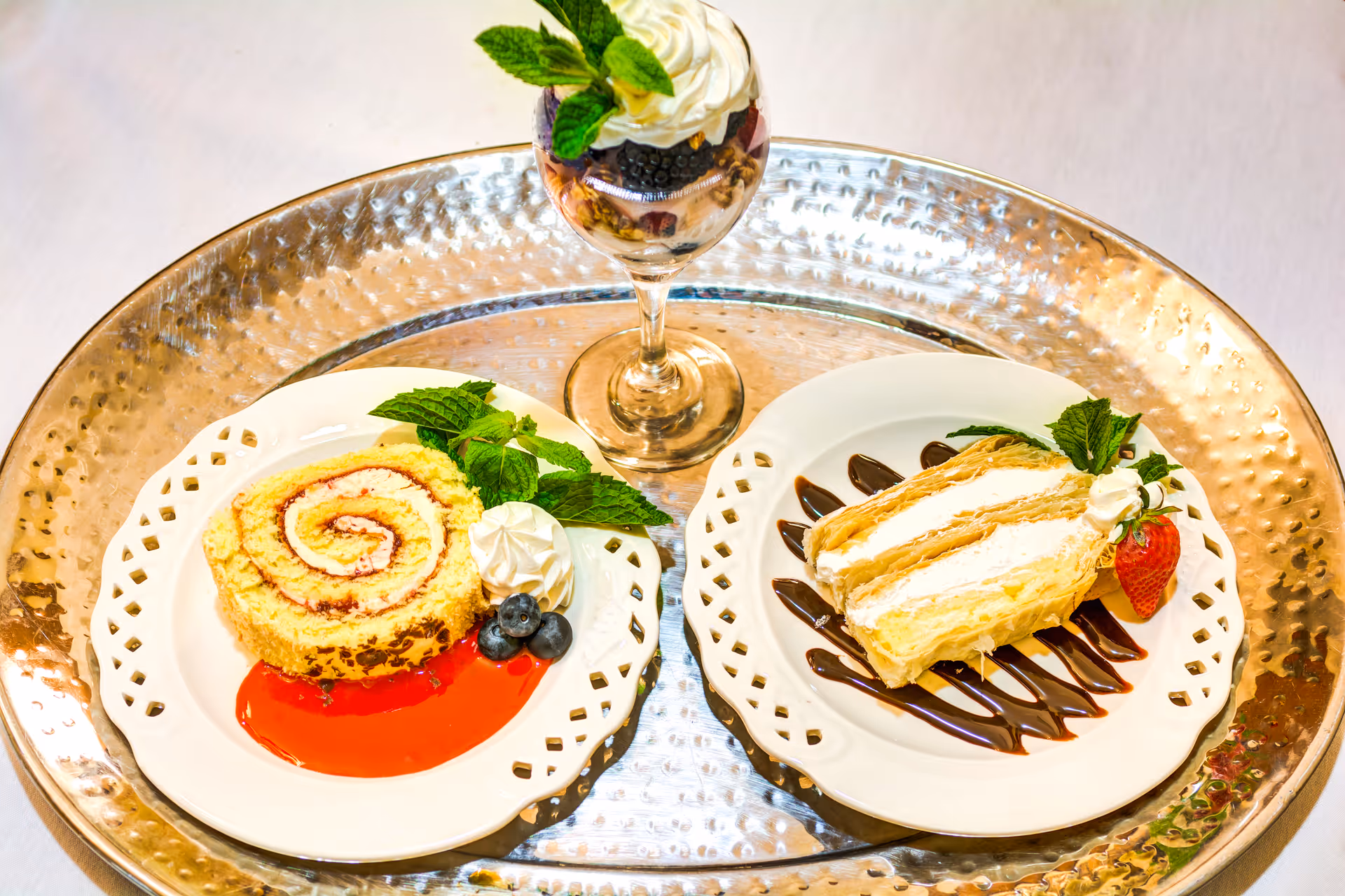 A silver tray holding two white plates with desserts and a dessert glass. One plate has a rolled cake with cream, garnished with mint leaves, blueberries, and a dollop of whipped cream on a red sauce. The other plate has a slice of layered cream cake with chocolate drizzle, a strawberry, mint leaves, and whipped cream. The dessert glass contains layered berries, granola, and whipped cream, topped with mint leaves.