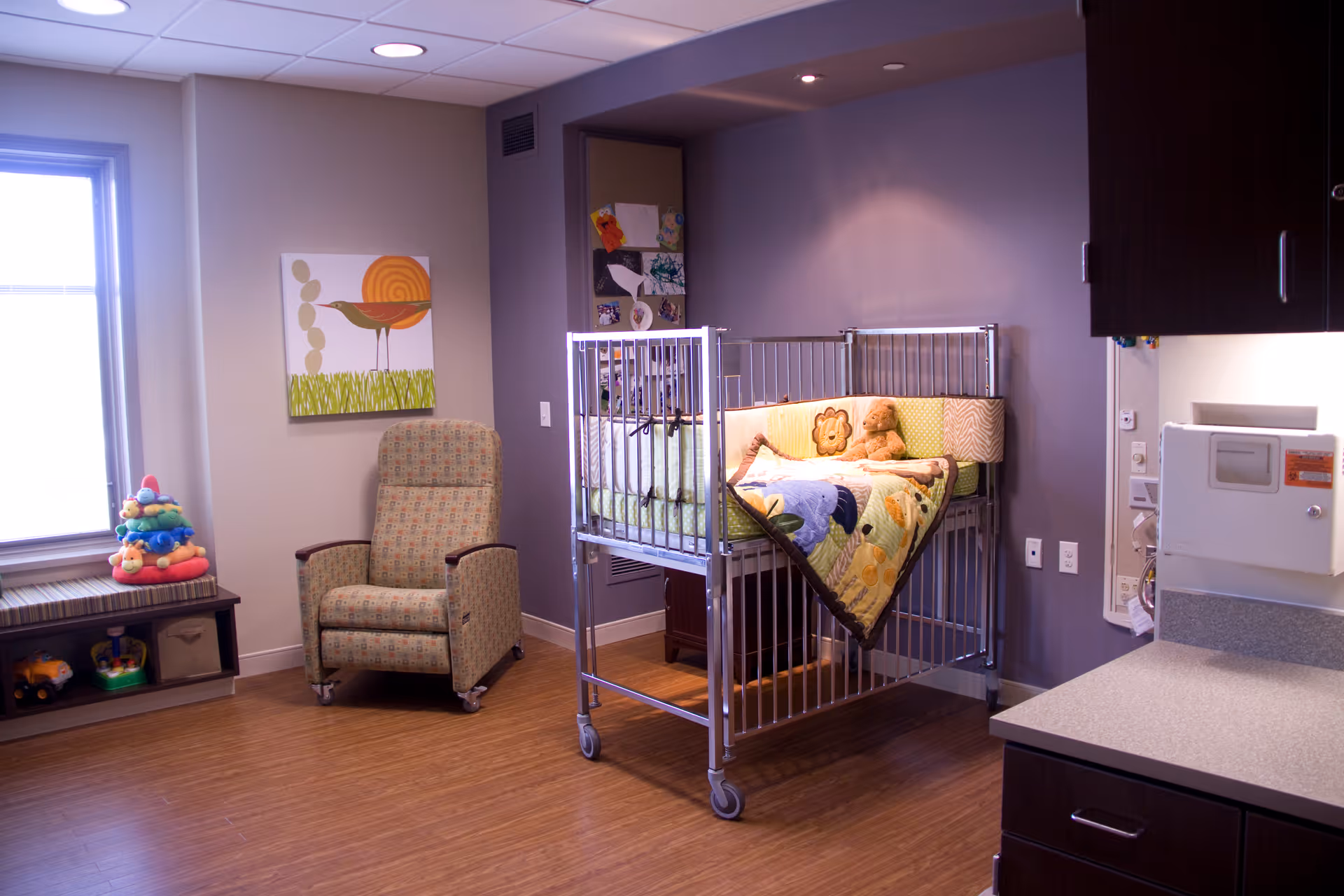 Cozy interior room with a wheeled metal crib draped with a colorful quilt, a patterned recliner, toys on a low shelf, and wall art.