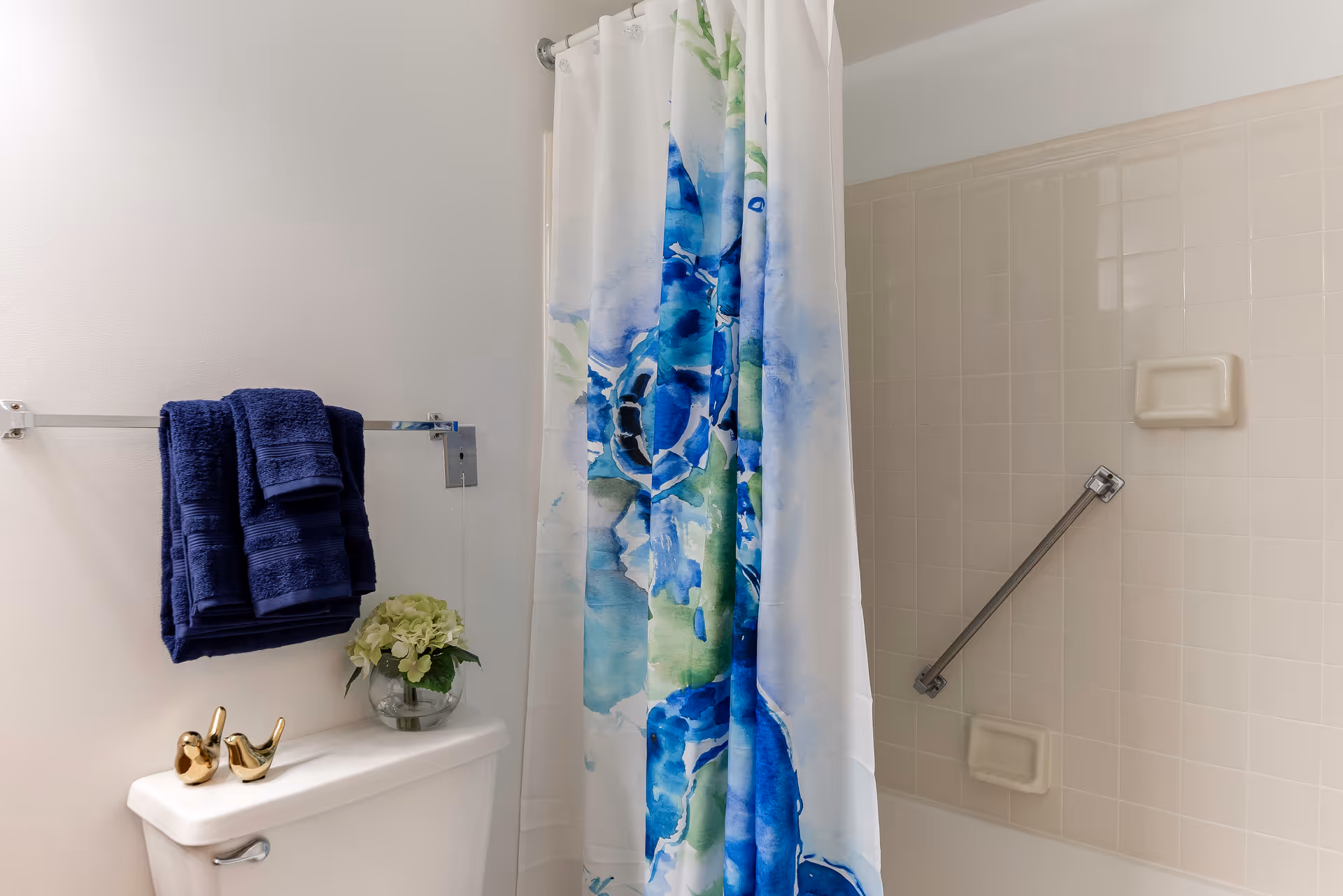 A bathroom with a white toilet topped with two small gold bird figurines and a glass vase with green flowers. Above the toilet is a towel rack holding three folded dark blue towels. To the right is a shower area with beige tiled walls, a silver grab bar, soap holders, and a shower curtain featuring blue and green floral patterns.