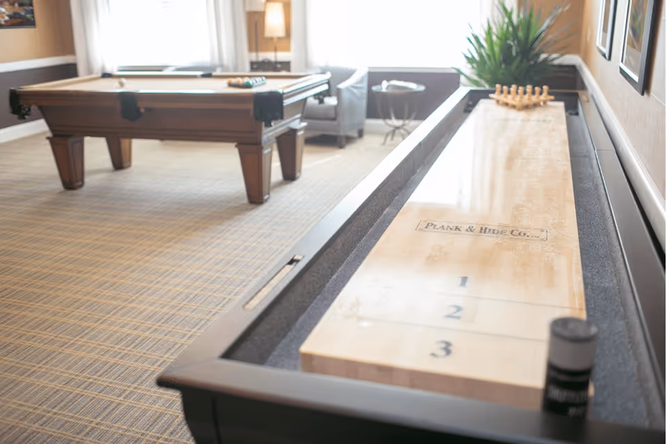 A recreational room featuring a shuffleboard table in the foreground and a pool table in the background. The room has carpeted flooring, a window with white curtains, a gray armchair, a small round side table, and a potted plant on a shelf along the wall.