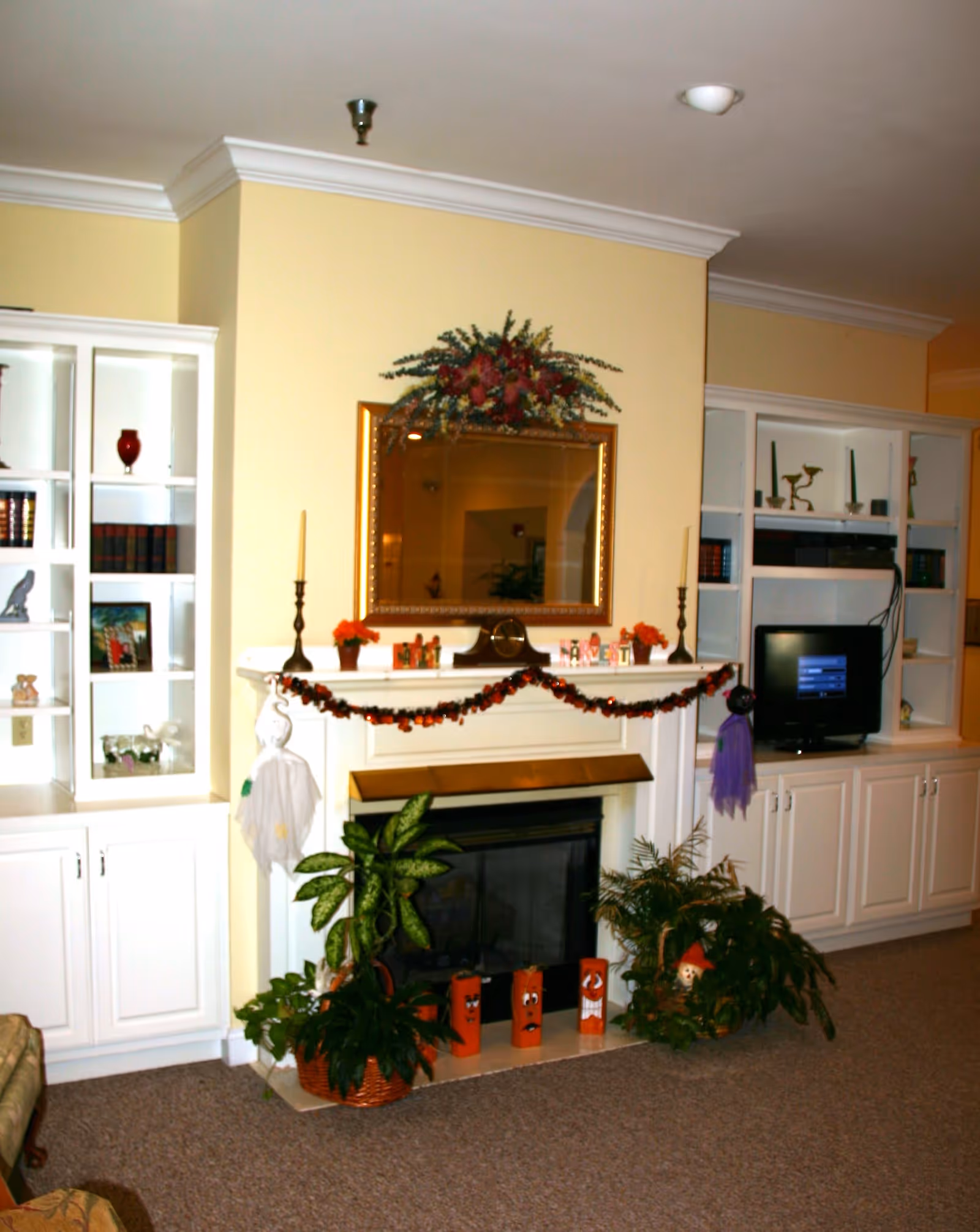 A cozy living room area with a white fireplace decorated with Halloween-themed garlands and small decorations. Above the fireplace is a large gold-framed mirror with a floral arrangement on top. On either side of the fireplace are white built-in shelves and cabinets, one side holding a small TV and various decorative items. There are several green potted plants placed around the fireplace on the carpeted floor.