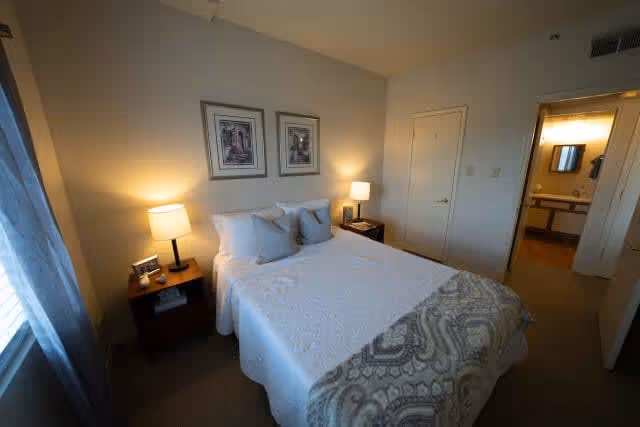A cozy bedroom with a neatly made bed featuring white bedding and a patterned throw blanket. There are two bedside tables each with a lamp, and two framed pictures hanging on the wall above the bed. A door leads to an adjacent bathroom with a visible sink and mirror.