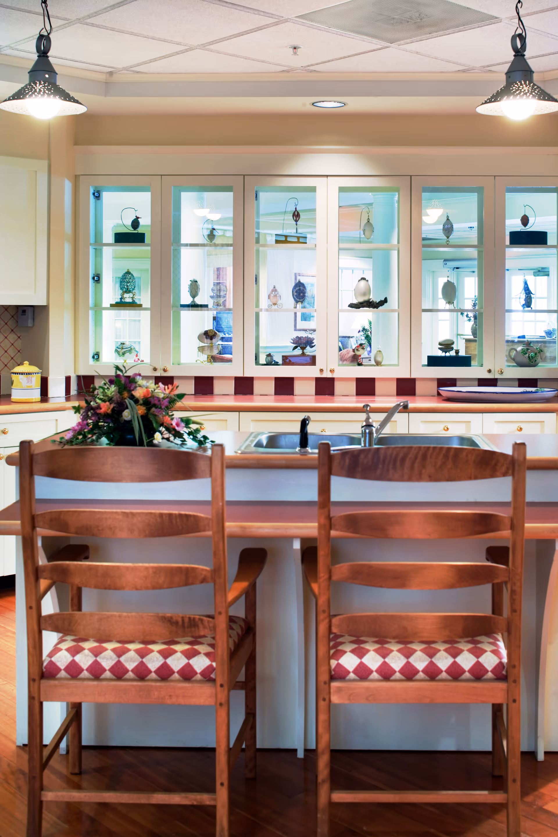 View of a kitchen area with two wooden chairs with red and white checkered cushions placed at a kitchen island. The island has a double sink with a faucet and a floral arrangement. Behind the island, there are white cabinets with glass doors displaying decorative items. Two pendant lights hang from the ceiling above the island.