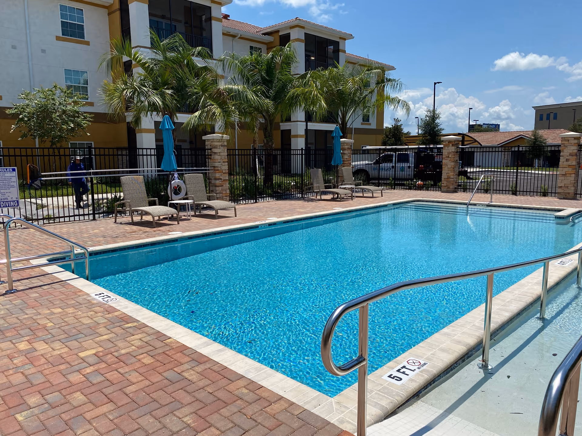 Outdoor swimming pool with clear blue water surrounded by a brick patio. Several lounge chairs and closed blue umbrellas are placed along the poolside. Palm trees and a multi-story building are visible in the background under a clear blue sky.