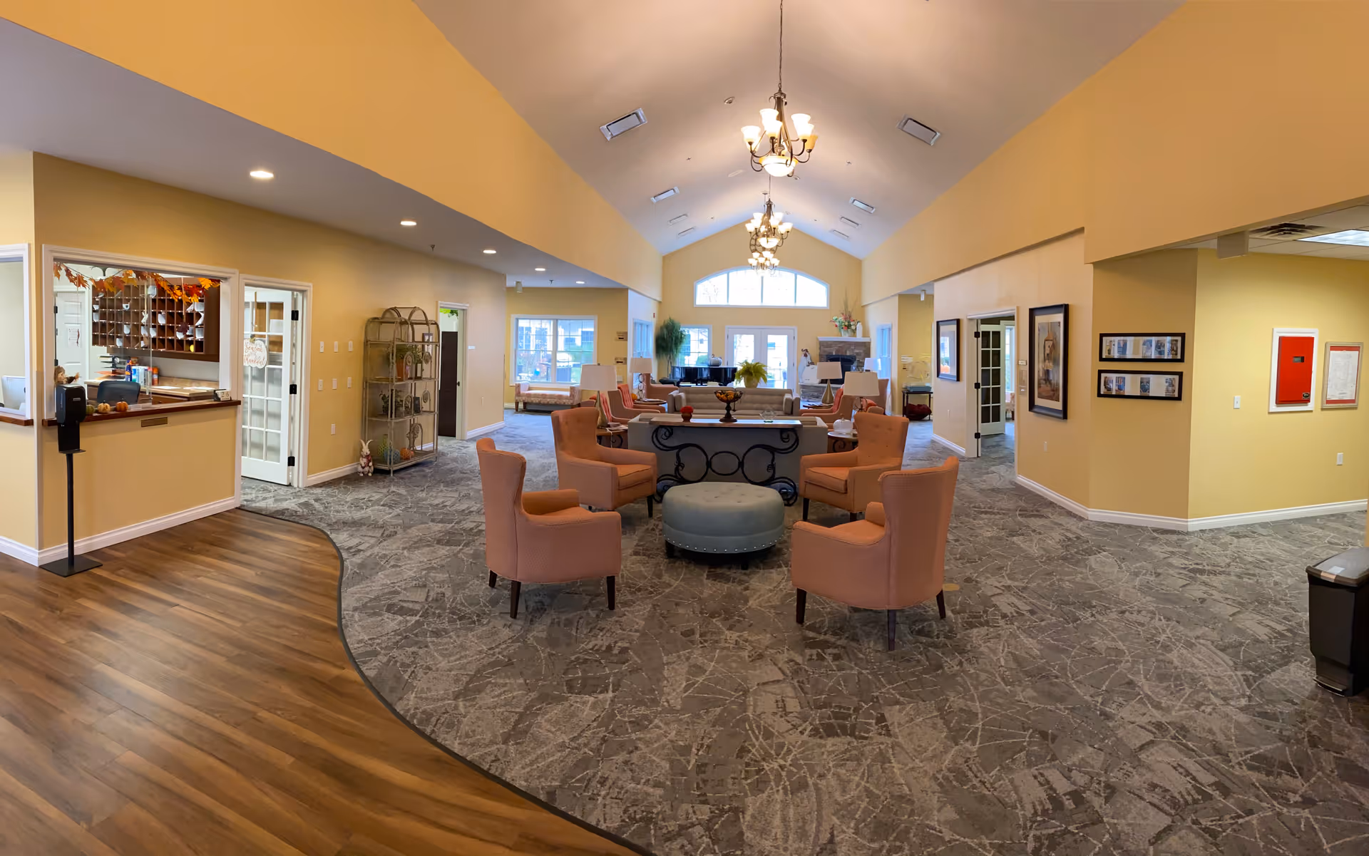 Spacious and well-lit assisted living community common area with high vaulted ceilings, multiple chandeliers, and a mix of carpet and wood flooring. The room features several comfortable armchairs arranged around a central ottoman, a reception desk on the left, large windows at the far end, and framed artwork on the walls.