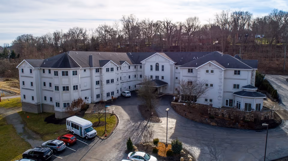 Three-story white senior living building with a circular driveway, parked cars and a shuttle van, set against leafless trees.