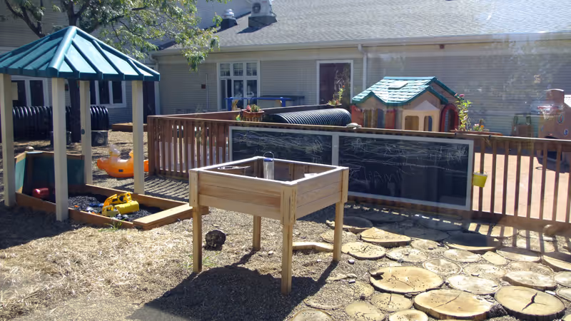 Outdoor play area with a wooden sandbox, a small playhouse, a chalkboard fence, a covered sandbox, and various toys on a ground covered with mulch and wooden stepping stones. A building with windows is visible in the background.