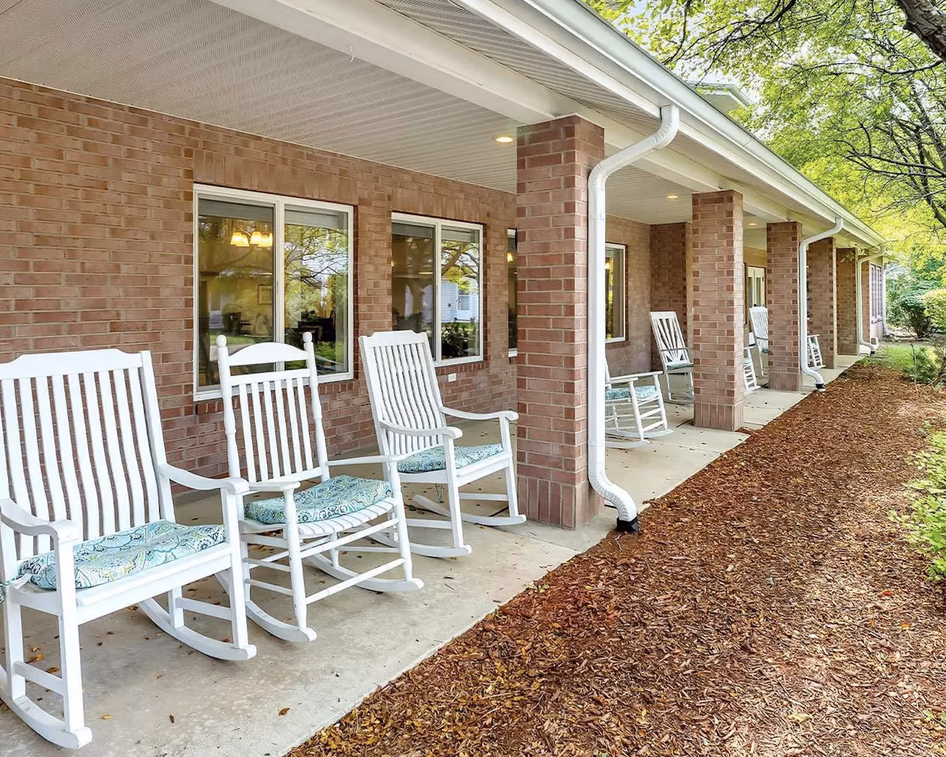 A covered outdoor patio area with several white wooden rocking chairs with patterned cushions lined up against a brick wall. The patio has a concrete floor and is supported by brick columns. There are windows on the brick wall and trees and greenery visible in the background.