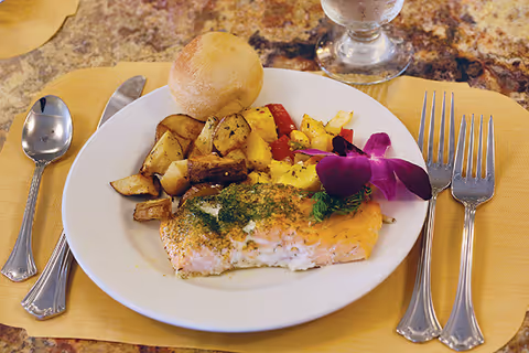 A plate of food on a table with a piece of cooked salmon garnished with herbs and an edible purple flower, roasted potatoes, mixed vegetables, and a bread roll. The plate is set on a yellow placemat with a spoon, fork, and knife arranged around it, and a glass of water in the background.