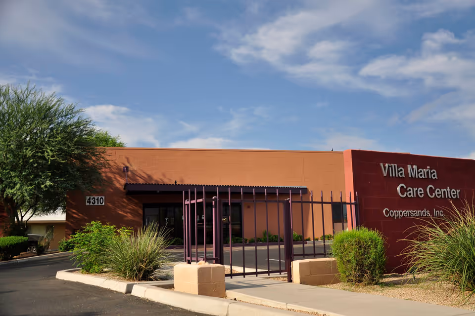 Exterior view of Villa Maria Care Center building with a clear blue sky, a gated entrance, and surrounding greenery including bushes and a tree.