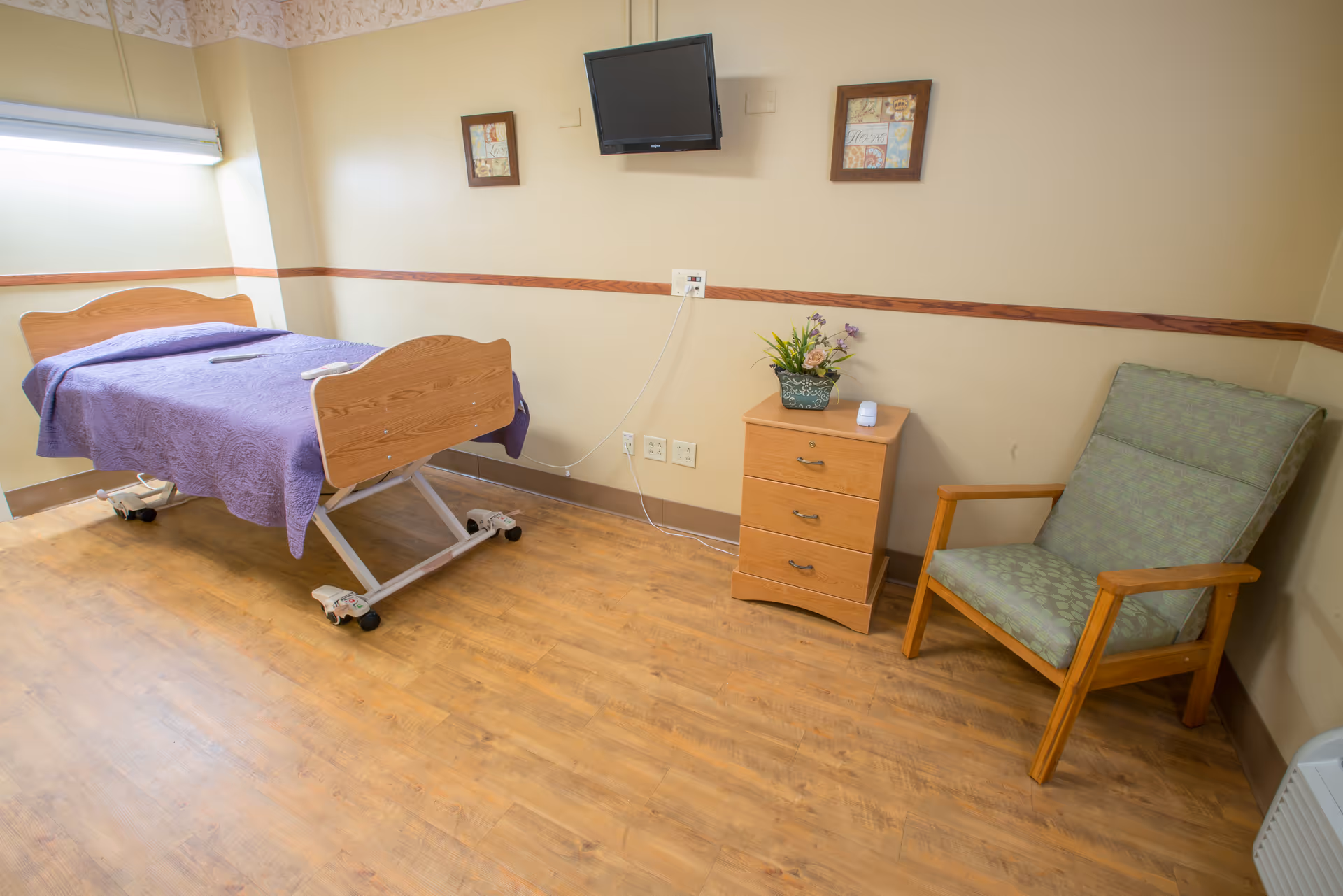 A nursing facility patient room with an adjustable bed covered by a purple blanket, a wooden nightstand topped with flowers, a green armchair, and a wall-mounted TV.