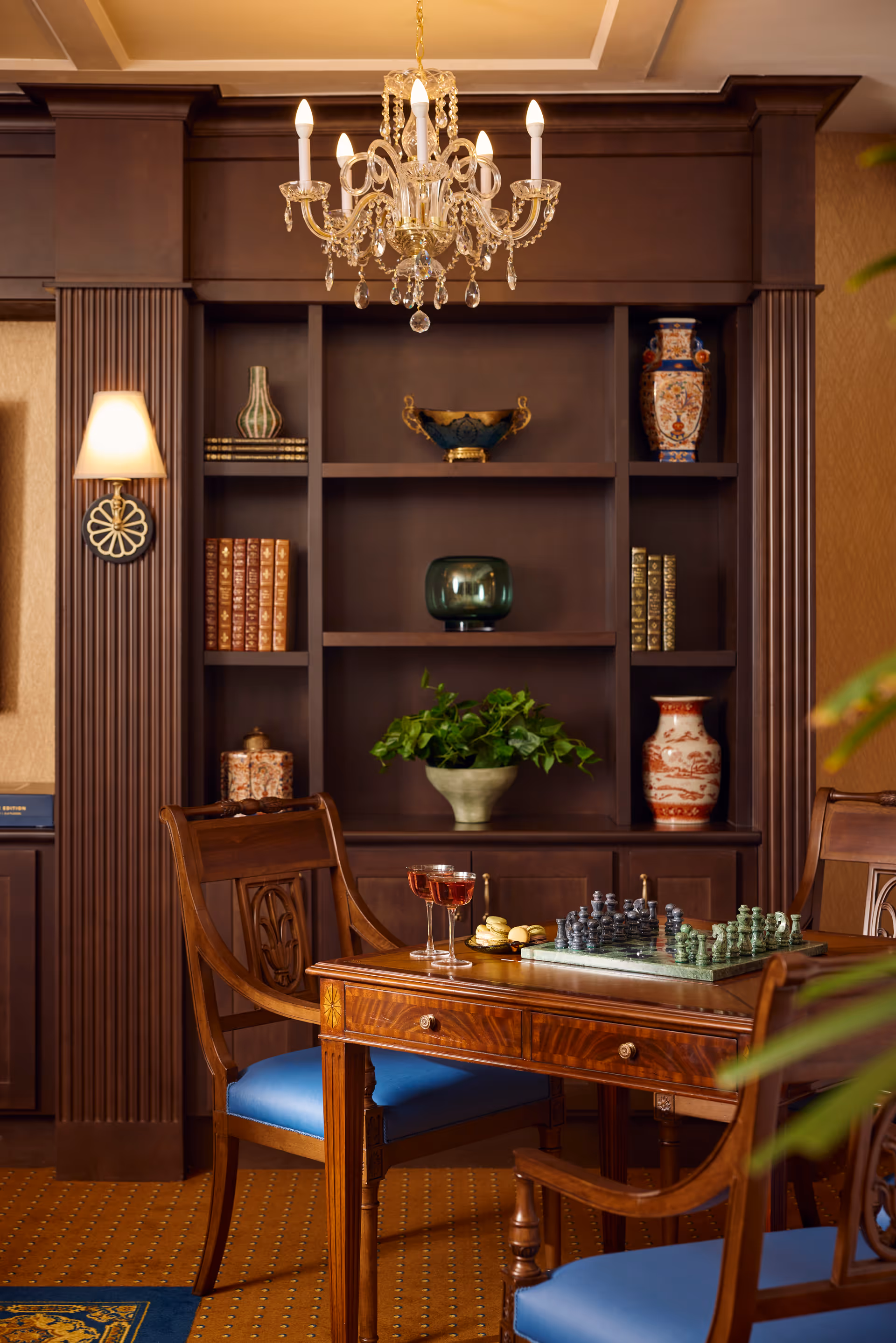A cozy interior room with a wooden table and two chairs with blue cushions. On the table, there is a chessboard with chess pieces, two glasses of red wine, and some snacks. Behind the table is a dark wooden built-in bookshelf with decorative items including vases, books, and a plant. A chandelier hangs from the ceiling, and a wall sconce with a light is mounted on the left side of the bookshelf.