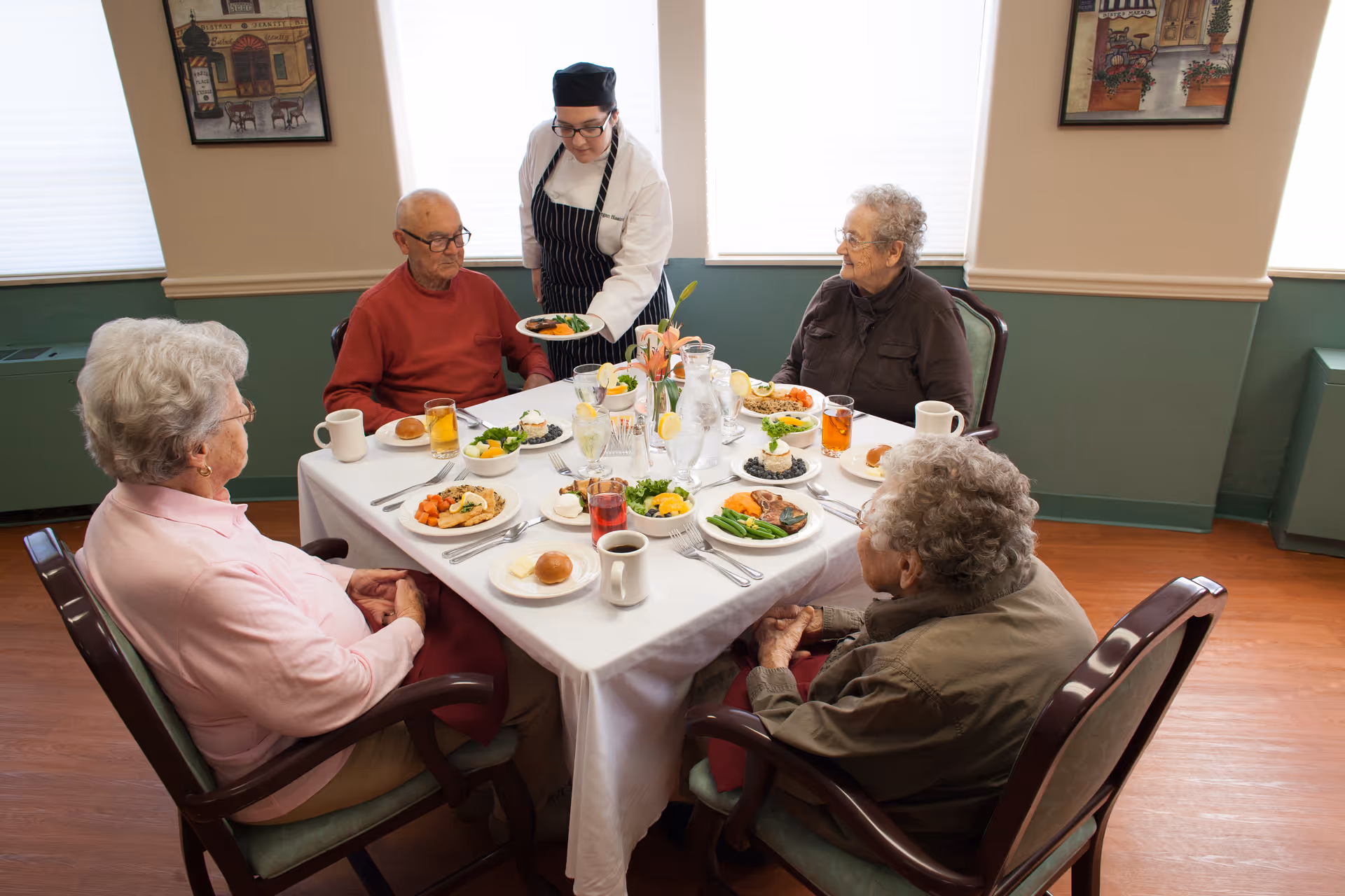 A server brings plates to four elderly residents seated around a table set with meals in a dining room.