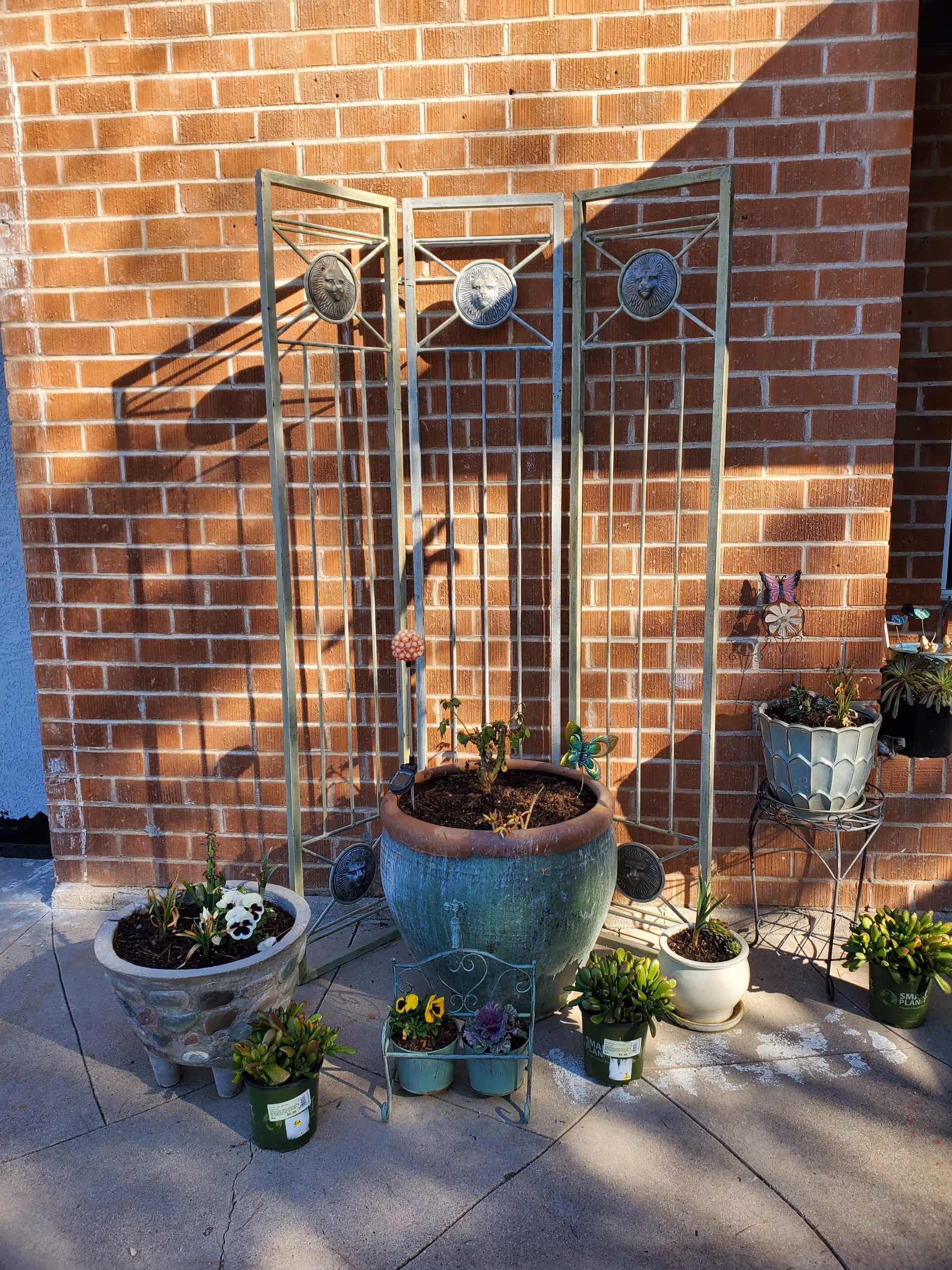 A small outdoor garden area with various potted plants arranged on a concrete surface against a red brick wall. There is a decorative metal trellis with lion head medallions behind a large green planter. Several smaller pots with flowers and succulents are placed around the large planter, some on the ground and some on a small metal stand.