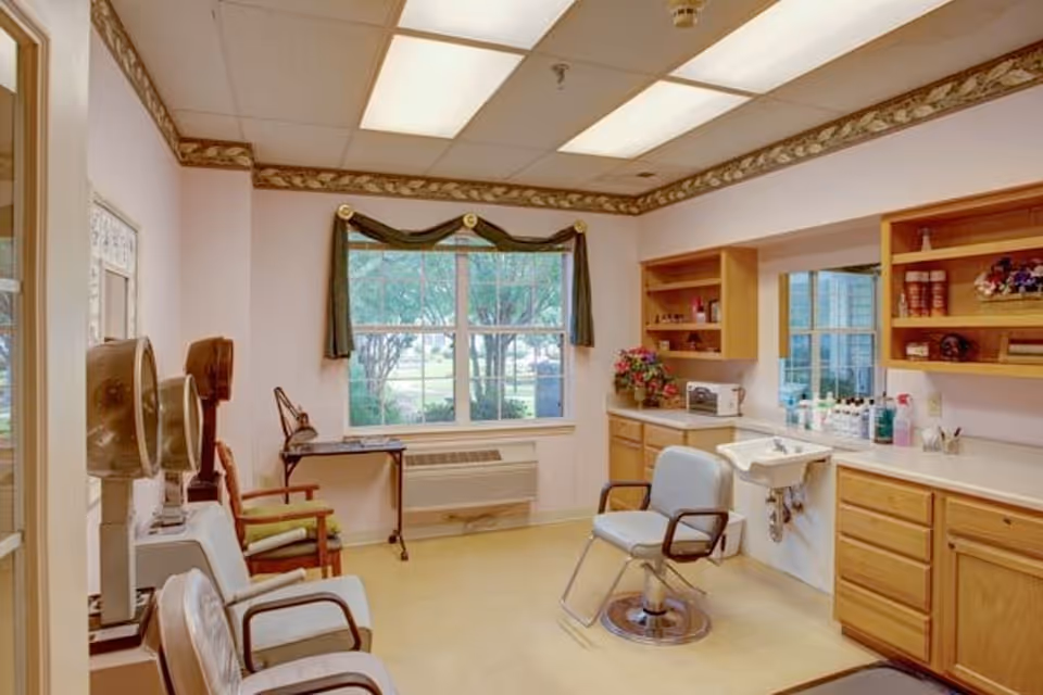 Interior view of a salon or grooming room in an assisted living facility. The room features salon chairs, hair dryers, a small sink, wooden cabinets with shelves holding various bottles and supplies, a window with a green valance, and a small table with a lamp. The walls are light-colored with a decorative border near the ceiling.
