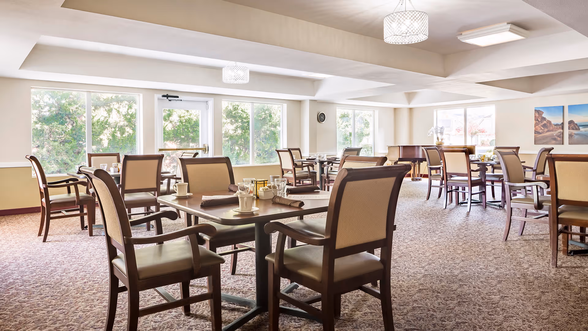 A bright dining room with multiple tables and chairs arranged neatly. Each table is set with cups, glasses, and napkins. Large windows allow natural light to fill the room, and a piano is visible in the background. The room has a carpeted floor and light-colored walls with framed artwork.