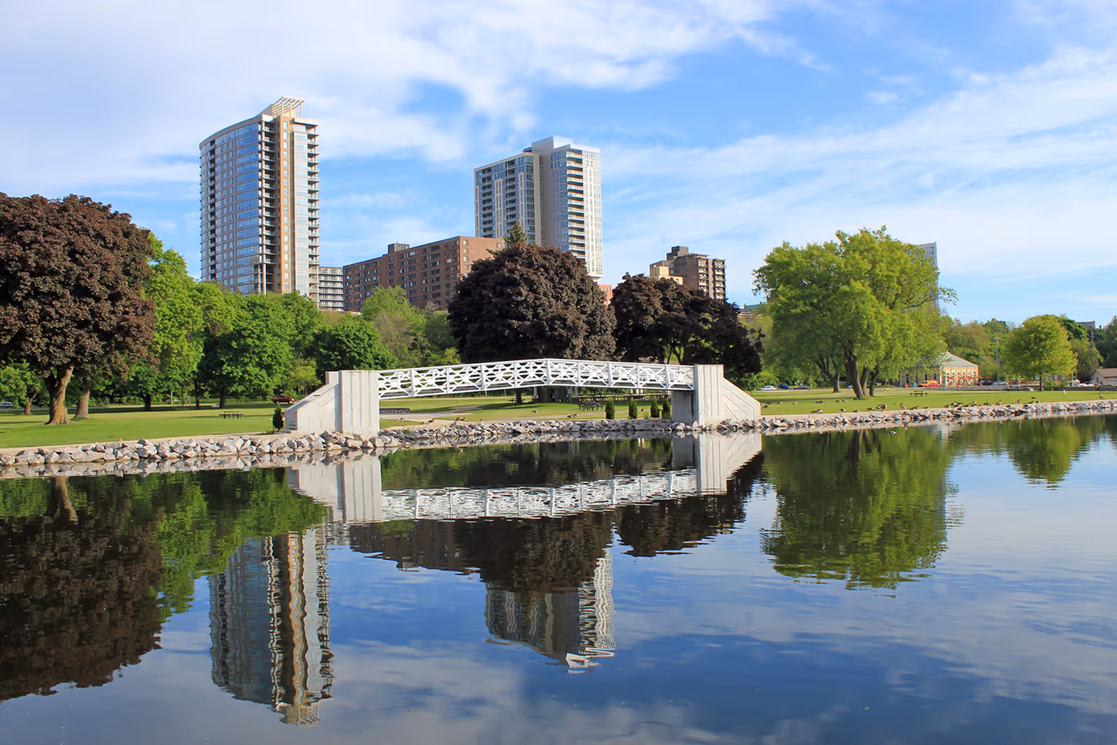 A serene outdoor scene featuring a small white pedestrian bridge over a calm body of water, with reflections of trees and tall buildings in the water. The area is lush with green trees and grass under a partly cloudy blue sky.