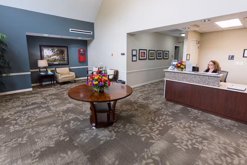 Reception area of Independence Village Of Midland with a wooden round table holding a colorful flower arrangement in the center. To the right, a receptionist sits behind a dark wood desk with a mosaic tile front. The walls are painted in neutral tones with framed pictures and a seating area with chairs and a side table with a lamp is visible on the left.