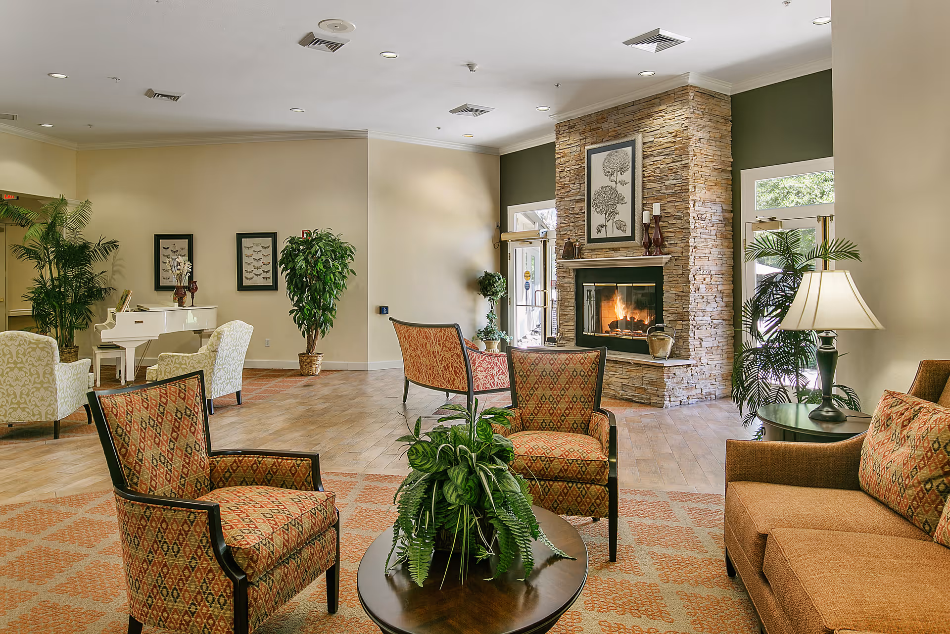 A comfortable common lounge with patterned armchairs, sofa and coffee table centered around a stone fireplace, with plants and a white piano in the background.