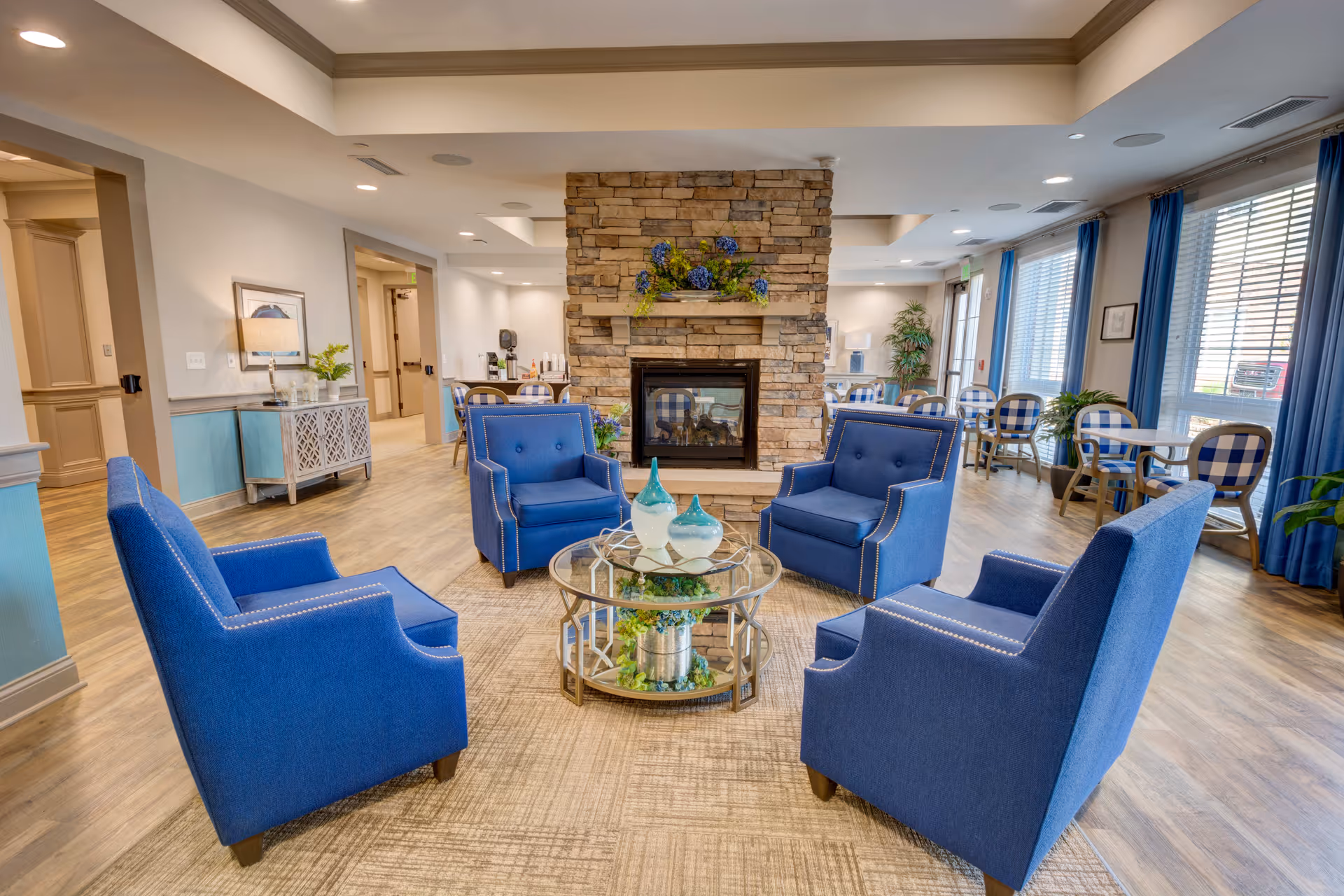 A cozy seating area in a senior living facility with four blue armchairs arranged around a glass coffee table. The table has decorative vases and greenery underneath. Behind the seating area is a stone fireplace with floral decorations on the mantel. The room has large windows with blue curtains, wooden flooring, and additional tables and chairs in the background.