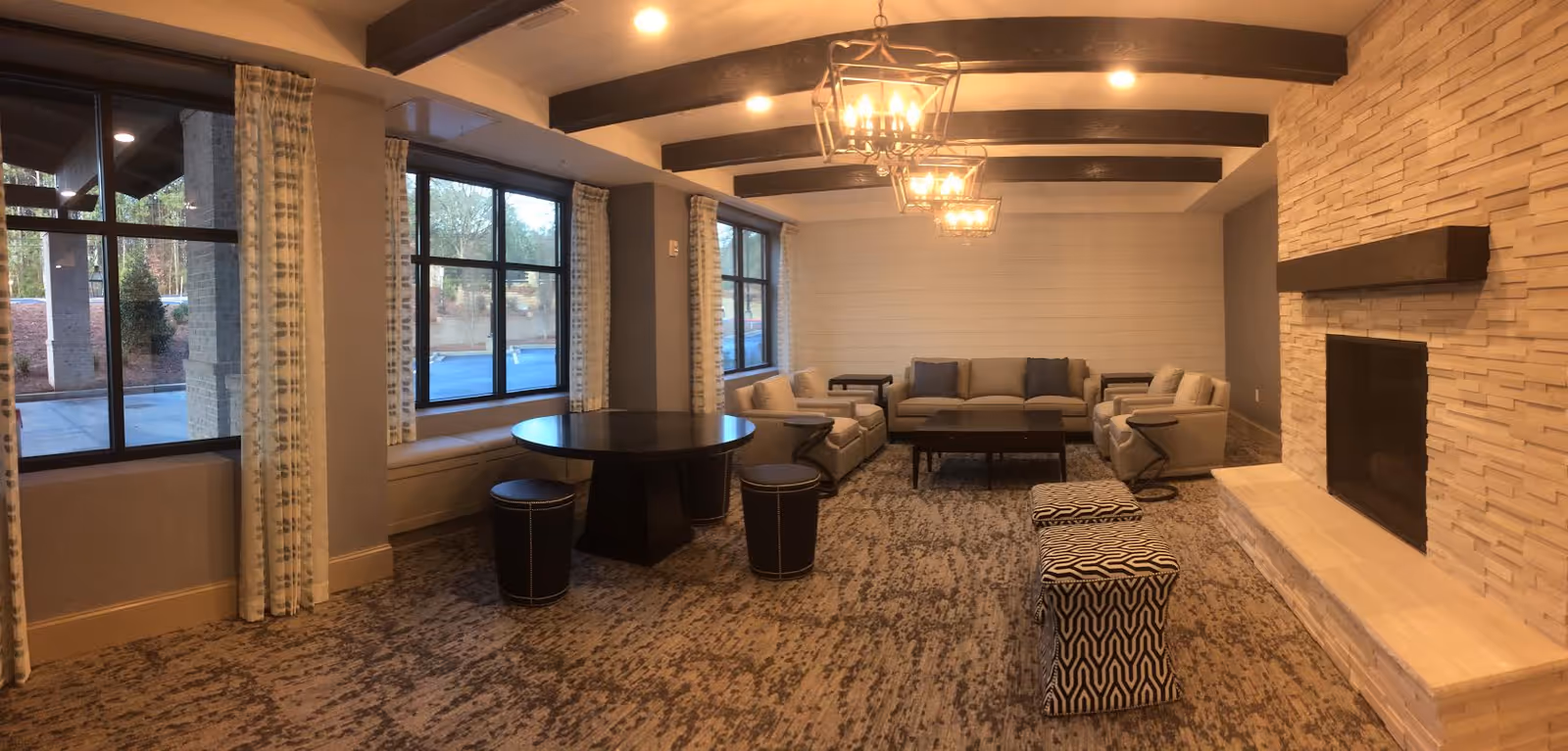 A cozy living room area with a stone fireplace on the right, two patterned ottomans in front of it, and a seating arrangement consisting of a sofa and armchairs with side tables. There is a round table with three stools near large windows with patterned curtains. The ceiling has exposed wooden beams and three hanging light fixtures.