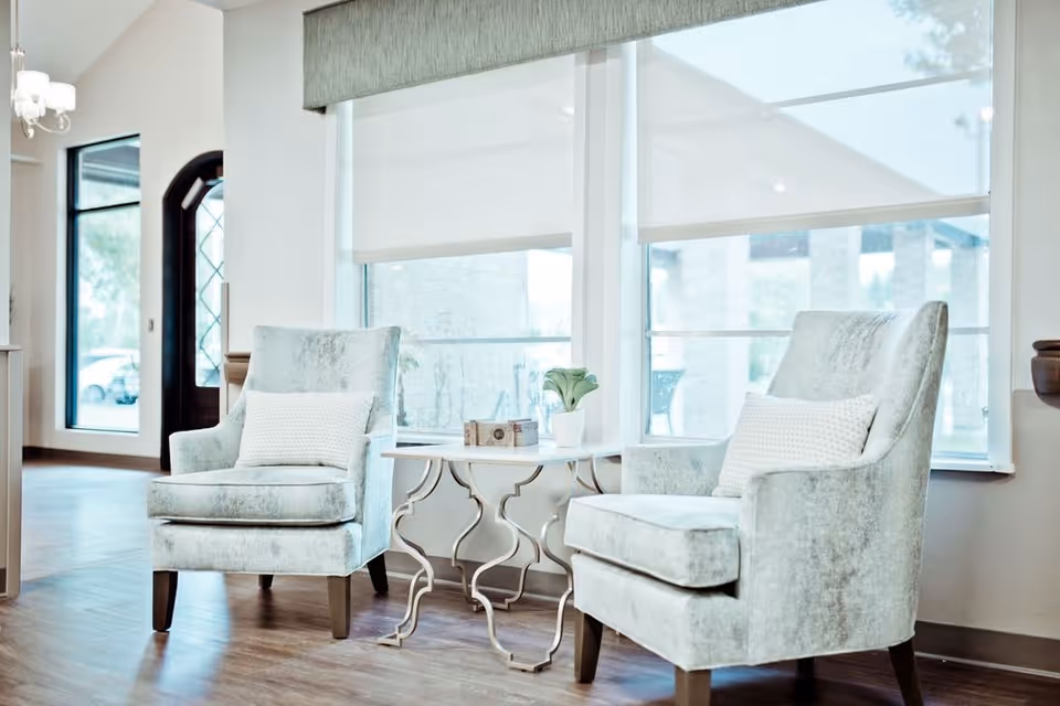 A bright and airy sitting area with two light gray upholstered armchairs, each with a white textured pillow. Between the chairs is a small white side table with a decorative plant and a small box. Large windows with white roller shades allow natural light to fill the space, and a dark wooden door with glass panels is visible in the background.