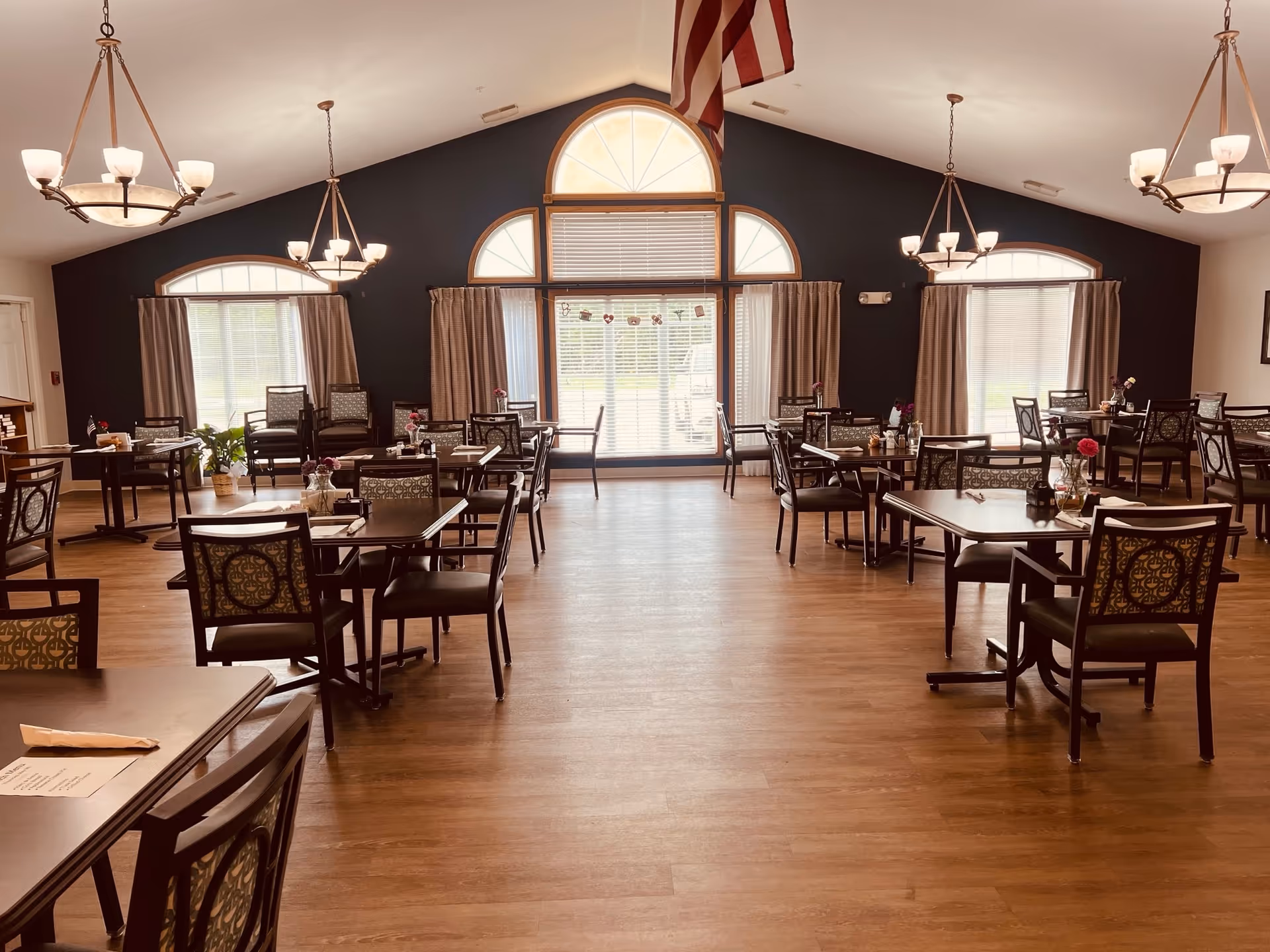 Spacious communal dining room with multiple tables and chairs, chandeliers, large arched windows, and wood flooring.