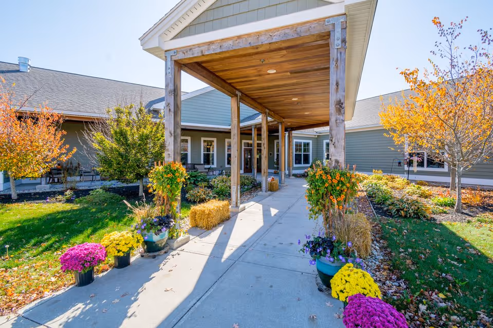 Entrance to Horizons Living & Rehab Center with a covered wooden walkway, colorful potted flowers, hay bales, and landscaped greenery with autumn trees under a clear blue sky.