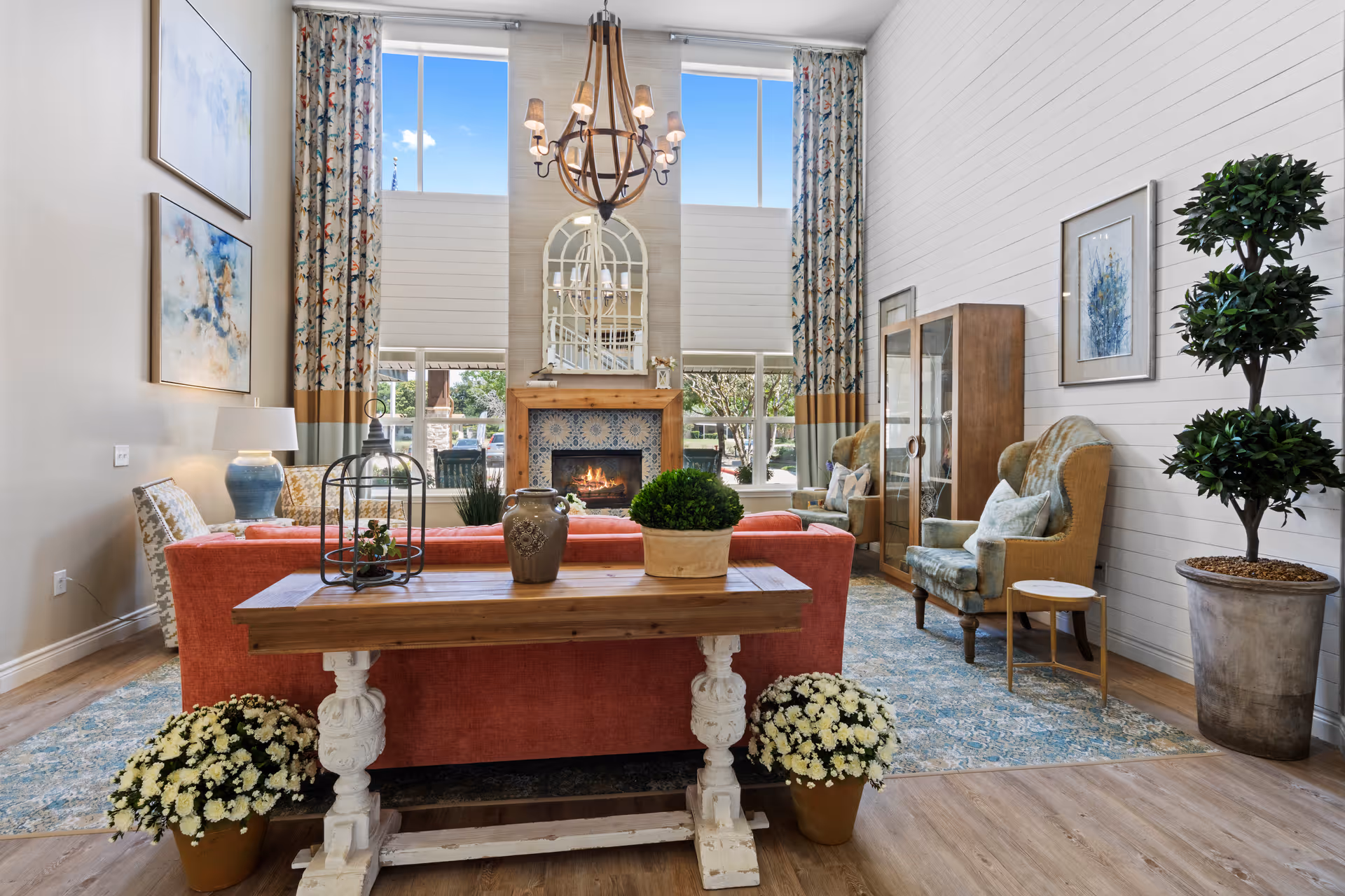 A bright and cozy living room with a red sofa facing a fireplace with a decorative tile surround. Above the fireplace is a large mirror and two tall windows with floral curtains. The room has wooden flooring, a blue patterned rug, two armchairs, a wooden cabinet, and various decorative plants and vases. A chandelier hangs from the ceiling.