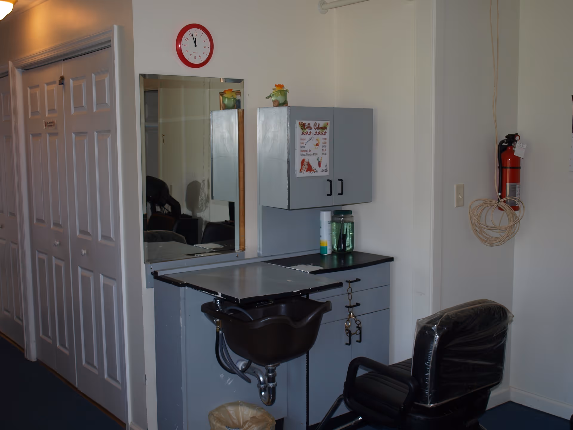 Interior view of a small hair salon area in a senior living facility. The image shows a black salon chair covered in plastic, a black wash basin attached to a gray cabinet with drawers, a mirror mounted on the wall, and a red clock above the mirror. There is a fire extinguisher mounted on the wall to the right and a coiled cable hanging next to it. The left side of the image shows white double closet doors.