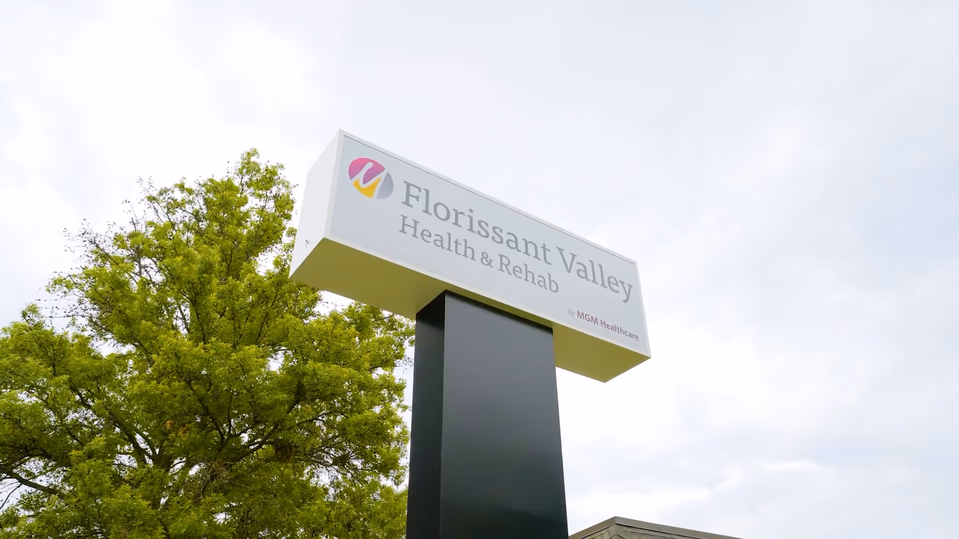 A tall roadside sign that reads "Florissant Valley Health & Rehab" next to a leafy tree under a cloudy sky.
