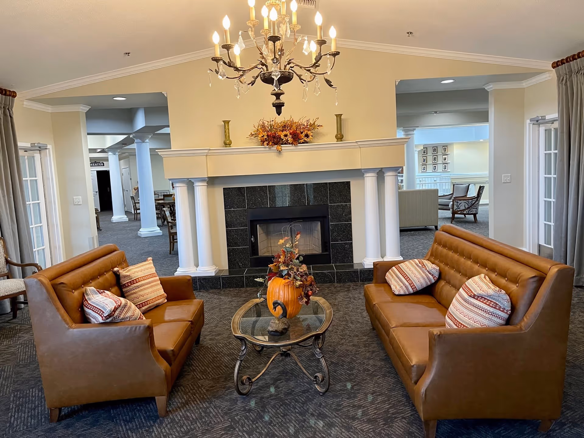 A cozy sitting area with two brown leather sofas facing each other, each with patterned pillows. Between them is a glass coffee table with a decorative pumpkin and autumn-themed floral arrangement. Behind the seating area is a fireplace with white columns on either side and a mantel decorated with vases and a fall floral arrangement. The room has beige walls, carpeted floors, and a chandelier hanging from the ceiling. In the background, there are open doorways leading to other rooms with additional seating and tables.