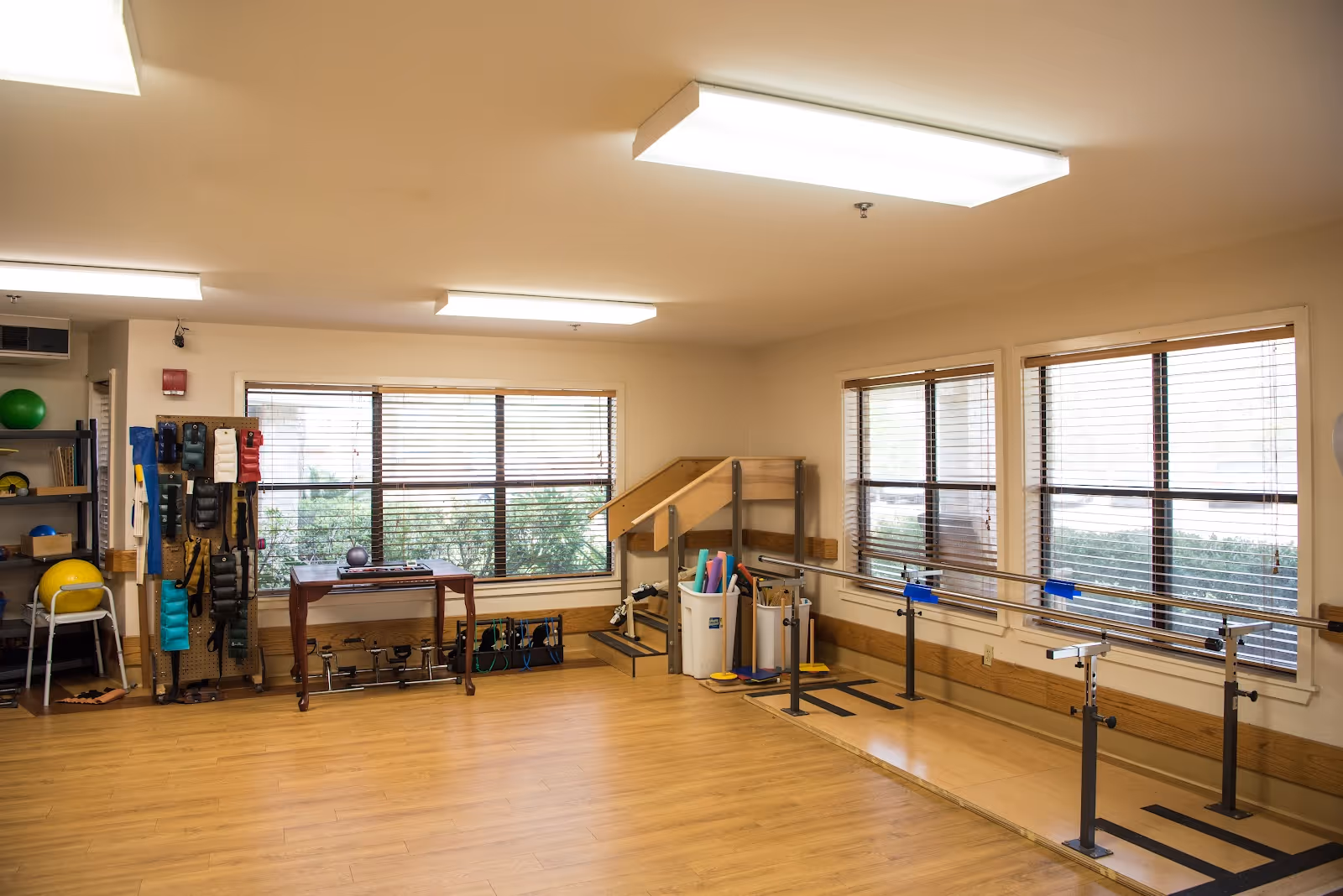 A spacious room with wooden flooring and large windows with blinds. The room contains physical therapy equipment including parallel bars, a small staircase with handrails, exercise bands, weights, and various therapy tools organized on shelves and racks. The room is well-lit with ceiling lights and has a clean, organized appearance.