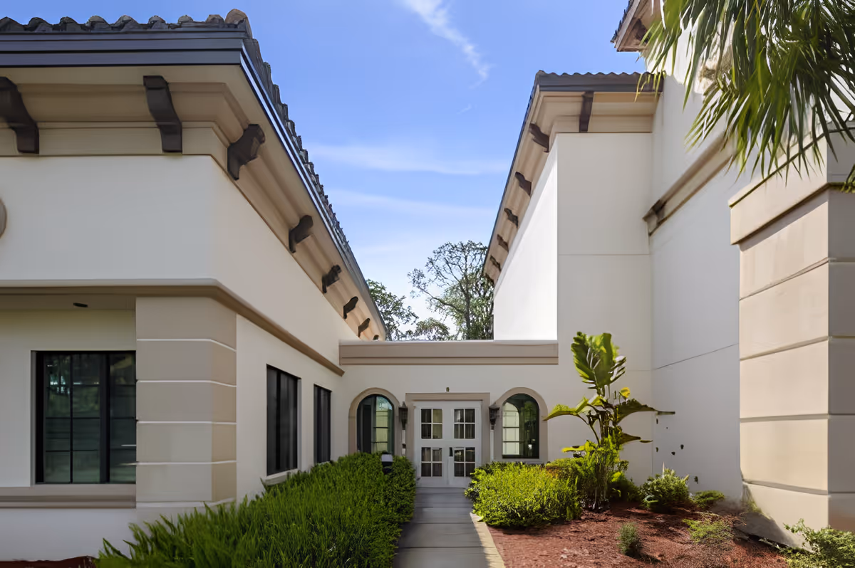 Pathway between two cream-colored building wings leading to a glass double-door entrance with landscaped shrubs and a palm.