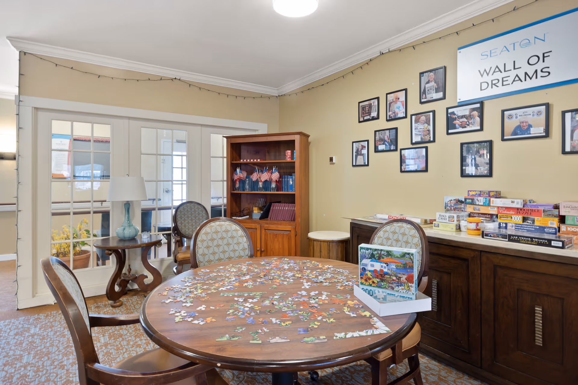 An activity/common room with a round table covered in jigsaw puzzle pieces, chairs, shelving, board games, and framed photos on the wall.