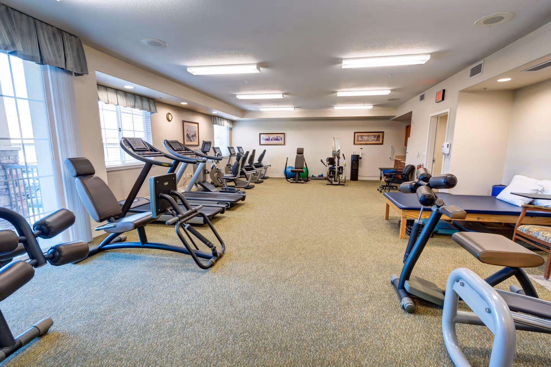 A spacious fitness room in a senior living facility with various exercise equipment including treadmills, stationary bikes, and strength training machines. The room has large windows with curtains allowing natural light, beige walls, and carpeted flooring. There are framed pictures on the walls and a therapy table with pillows on the right side.