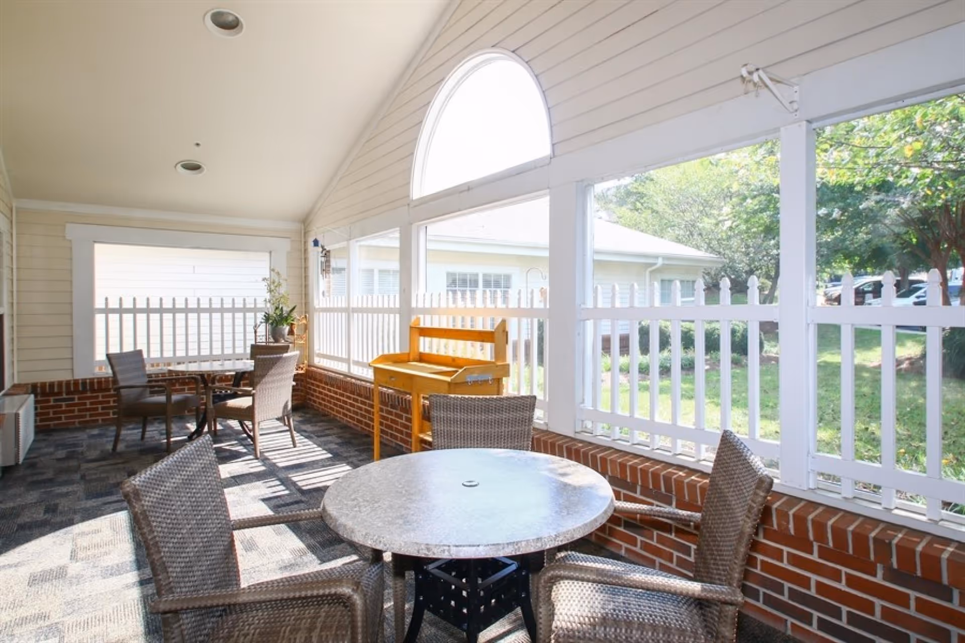 A bright enclosed patio area with a round table and four wicker chairs. The space has large windows with white picket fencing outside, brick lower walls, and a high ceiling with recessed lighting. There is a wooden bench and a small table with a plant in the background. Outside, green grass and trees are visible.
