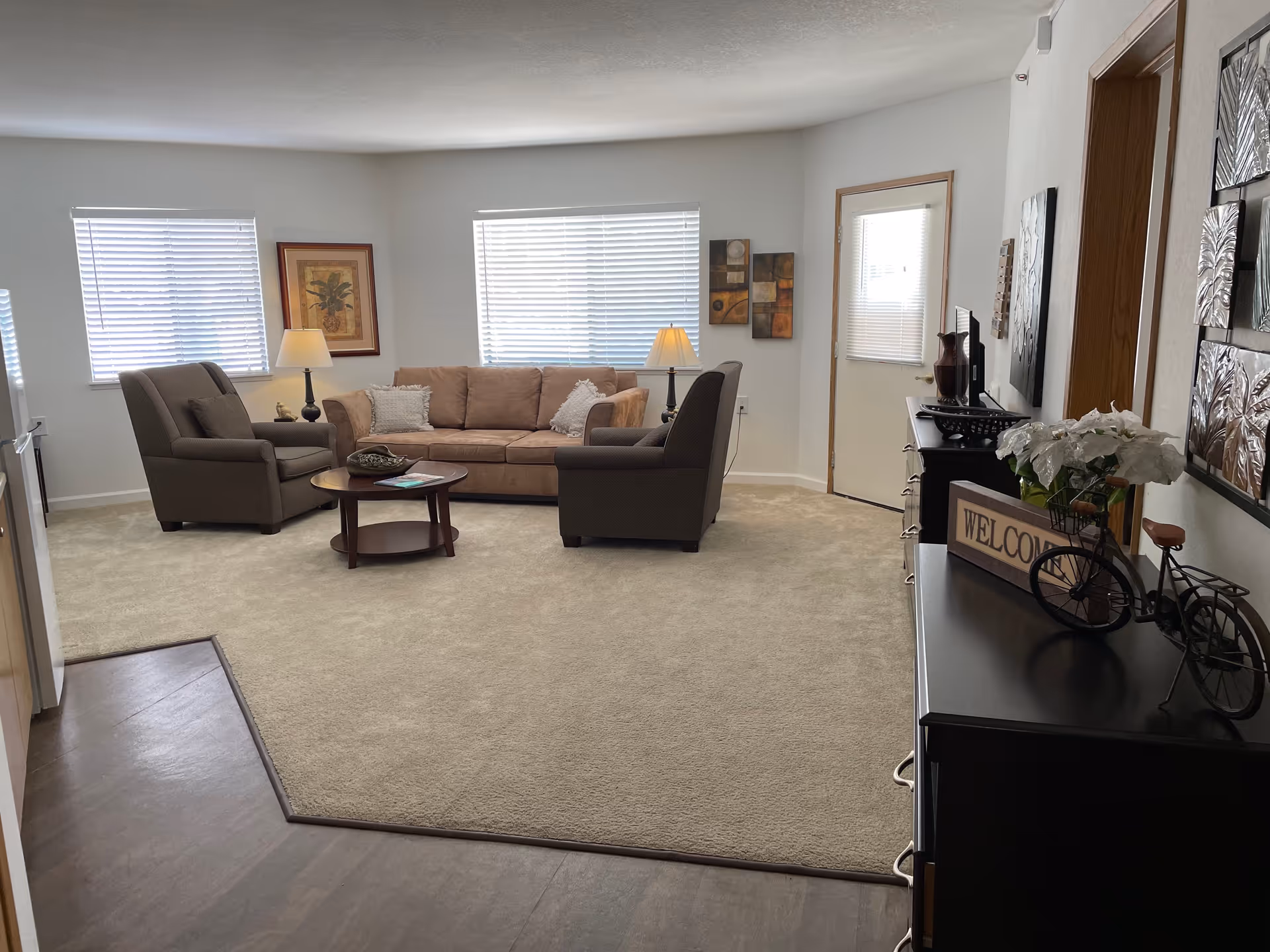 A cozy living room with beige carpet and light gray walls. The room features a beige sofa with two white pillows, two brown armchairs, a round wooden coffee table with decorative items, two table lamps, framed artwork on the walls, and a black dresser with a decorative bicycle and a 'WELCOME' sign. There are two windows with blinds and a door with a window blind.