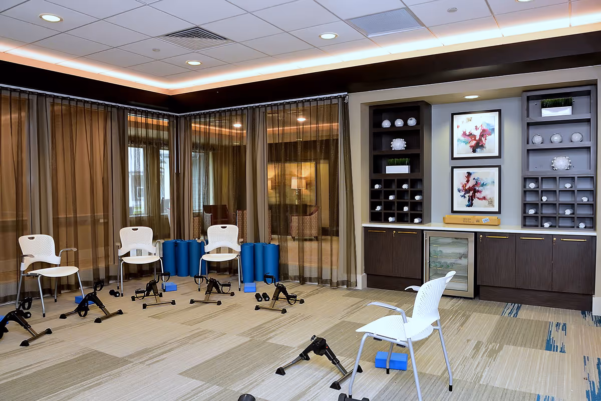 Bright activity/exercise room with chairs, multiple pedal exercisers and foam rollers, and built-in cabinets with artwork.