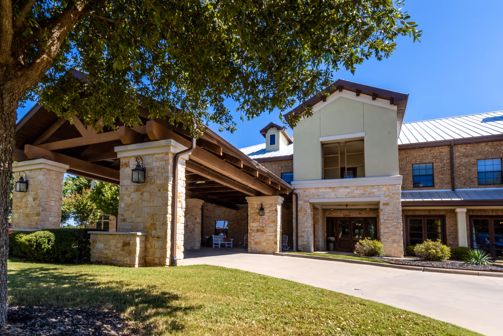 Front entrance and covered porte-cochere of a stone-and-brick assisted living building under a clear blue sky.