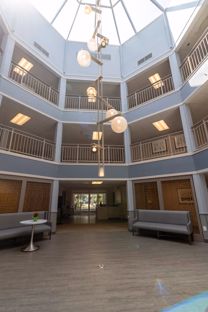 Interior view of a multi-story atrium with a large skylight ceiling and modern hanging light fixtures. The space features gray walls, railings on the upper floors, two gray sofas on either side, and a small round table with a potted plant. The area appears to be a lobby or common area with natural light coming through the skylight.